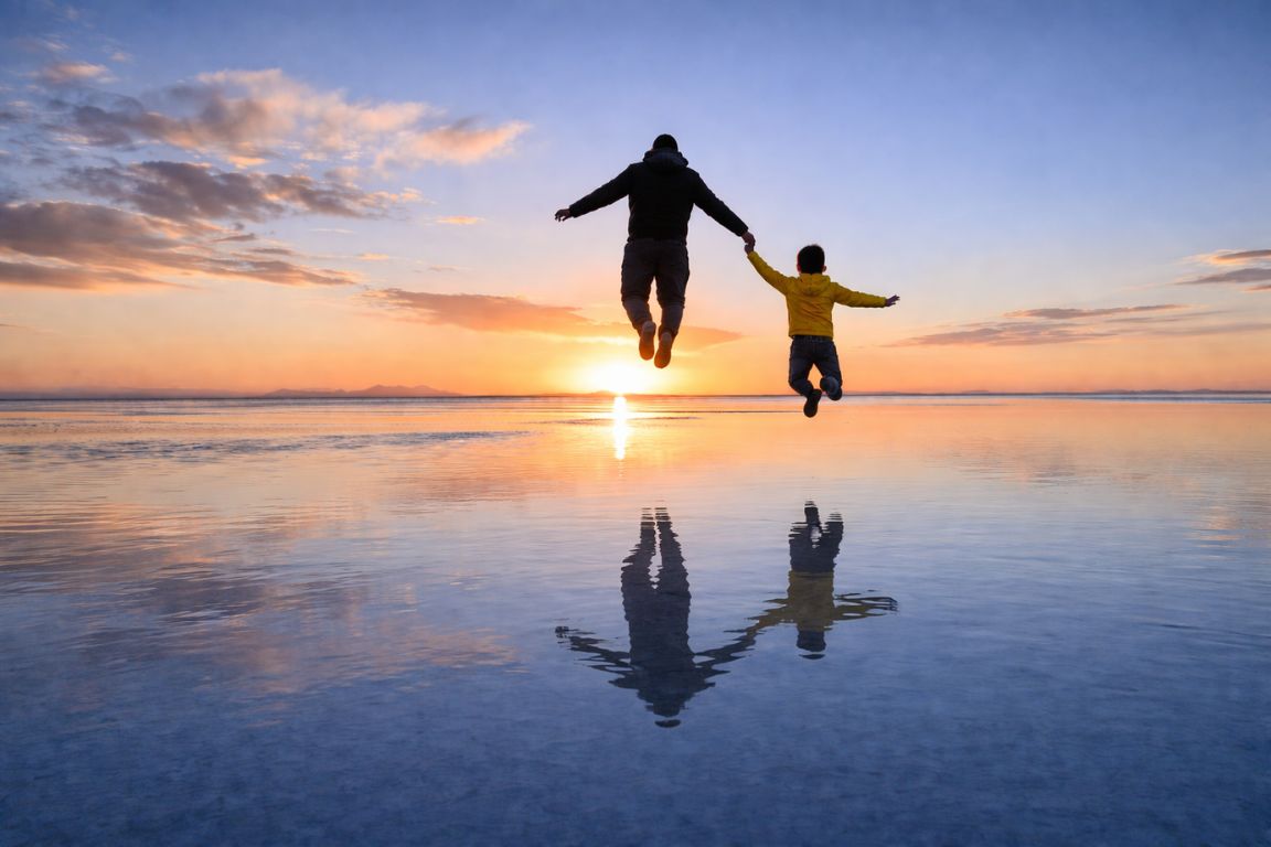 Iconic mirror effect photo of man and child jumping on Uyuni salt flats with colorful sunset sky, captured on a tour with Salar de Uyuni Tours