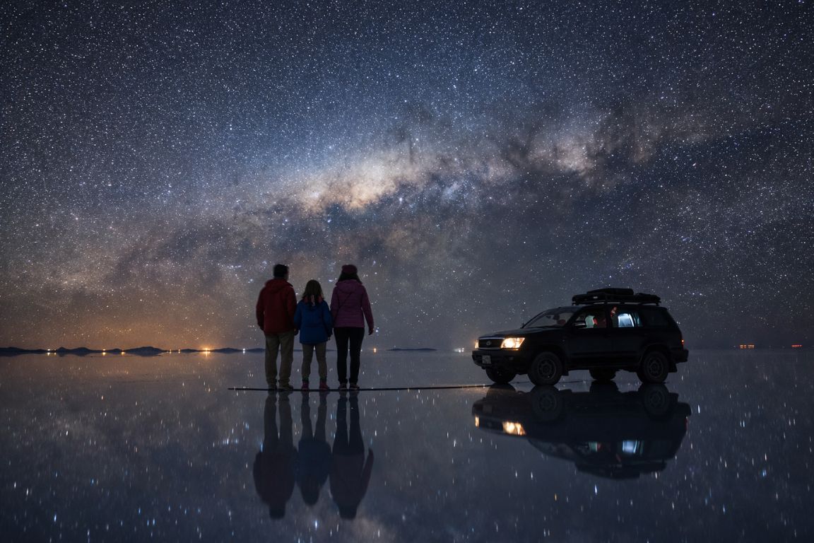 Night photography scene of Milky Way above Uyuni salt flats with reflections and tour vehicle, captured on a tour with Salar de Uyuni Tours